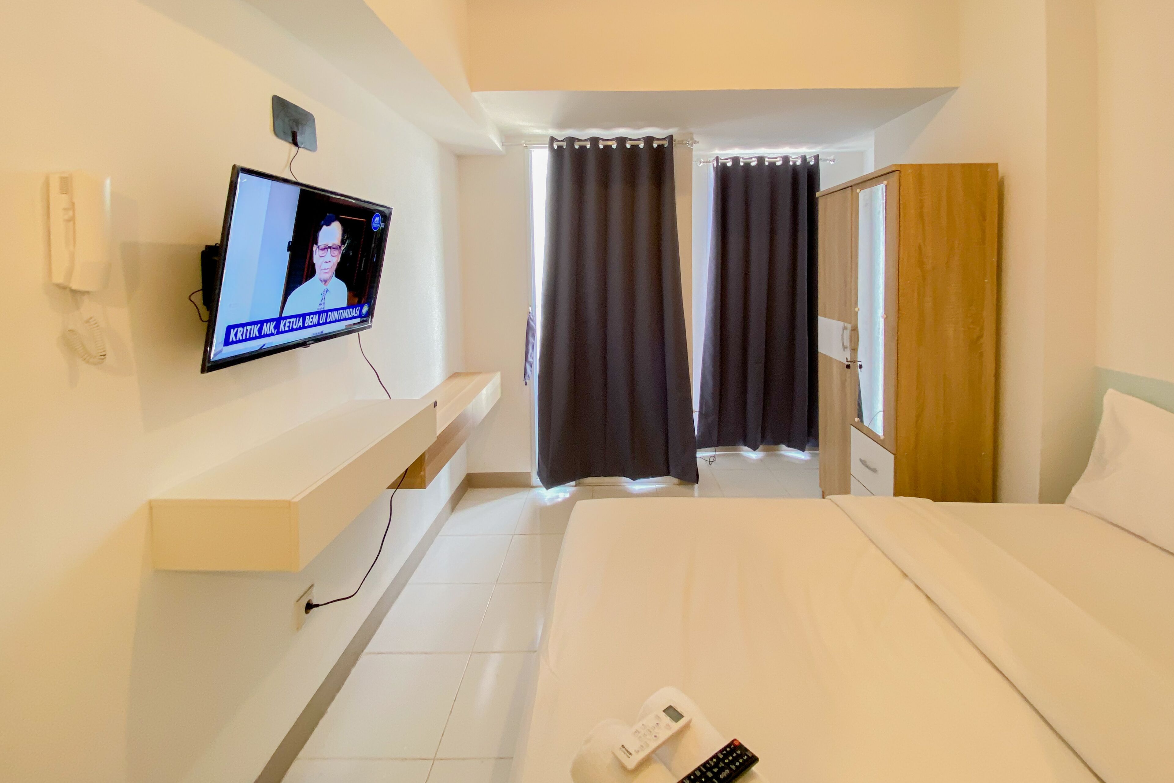 A hotel room with a bed, a TV mounted on the wall, a bedside table, a nightstand, and a window with curtains.