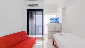 A hotel room with a red sofa, a bed with a white mattress, a television on a cabinet, and a sliding glass door leading to a balcony. - Cozy Stay Studio Apartment At M-Town Residence (Gading Serpong)