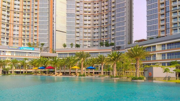 A large indoor swimming pool with clear blue water, surrounded by palm trees and lounge chairs. - Homey And Warm Studio At 28Th Floor Gold Coast Apartment (Jakarta)