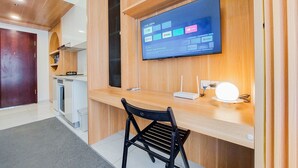 A modern room with a wooden desk, a flat-screen TV mounted on the wall, a black chair, and a small kitchenette area. - Minimalist Studio At Sky House Alam Sutera Apartment (Tangerang)