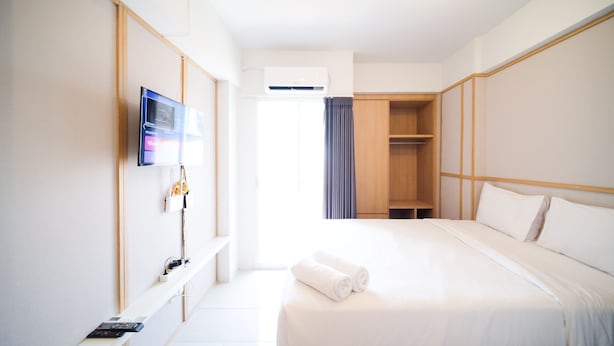 A hotel room with a bed, a TV mounted on the wall, a wardrobe, and a small table with a remote control. - Simple And Tidy Studio Apartment At Suncity Residence (Sidoarjo)