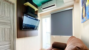 A living room with a brown sofa, a mounted flat-screen TV, an air conditioning unit, and a sliding door leading to a balcony. - Best Deal And Homey 1Br Apartment Grand Sentraland Karawang (Karawang)