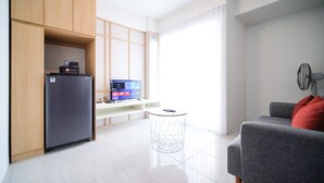 A modern living room with a grey sofa, a small round table, a TV on a stand, and a refrigerator. - Comfy And Spacey 2Br Apartment At Suncity Residence (Sidoarjo)