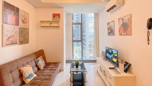 A living room with a brown sofa, a glass coffee table, and a TV on a white cabinet. There are paintings on the walls and a window with a view of buildings. - Comfort Stay 2Br At L'Avenue Pancoran Apartment (Jakarta)