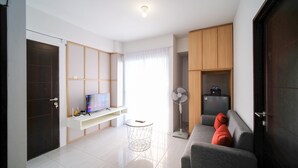 Uma sala de estar moderna com televisão de tela plana, ventilador de coluna, sofá cinza com almofadas vermelhas e geladeira compacta.