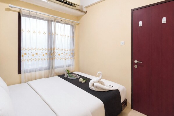 A hotel room with a bed, a window with sheer curtains, a door, and a small table with a towel and a flower.