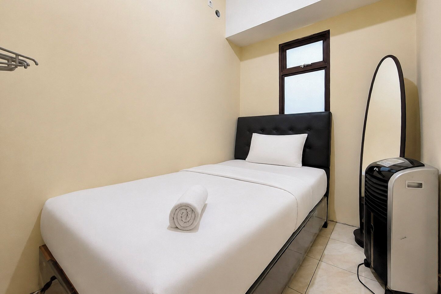 A single bed with a white mattress and a rolled white towel, a black headboard, a window with blinds, and a standing heater.