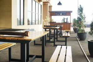 Een buitenzitplaats met houten tafels en banken, planten in potten en uitzicht op de straat.