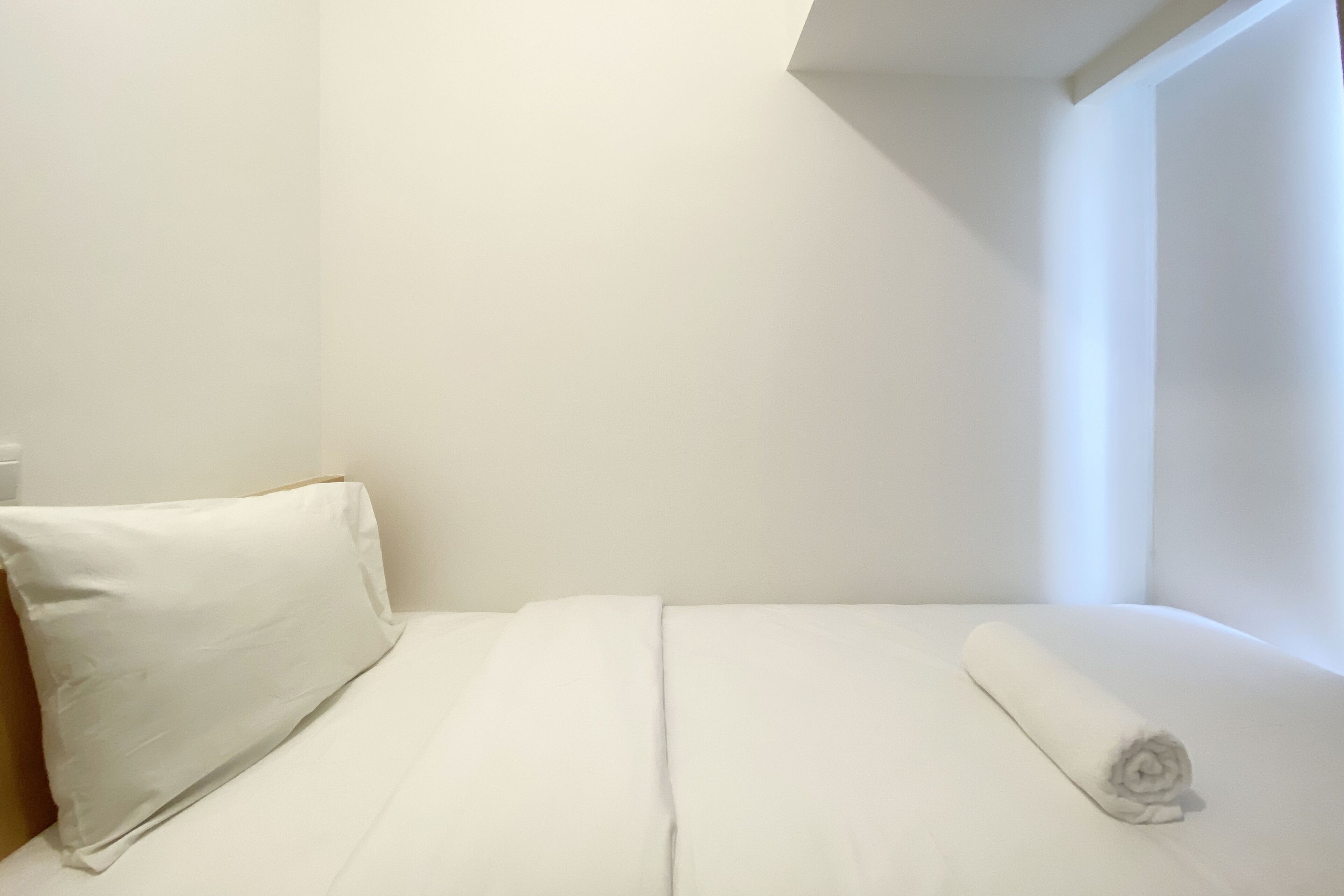 A neatly made bed with white linens and a rolled towel, in a room with plain white walls and a sloped ceiling.