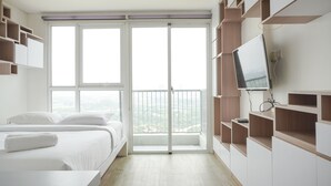 A modern bedroom with a large window, a bed, built-in shelving, and a TV. - Simply Look Studio At Casa De Parco Apartment (Pagedangan)