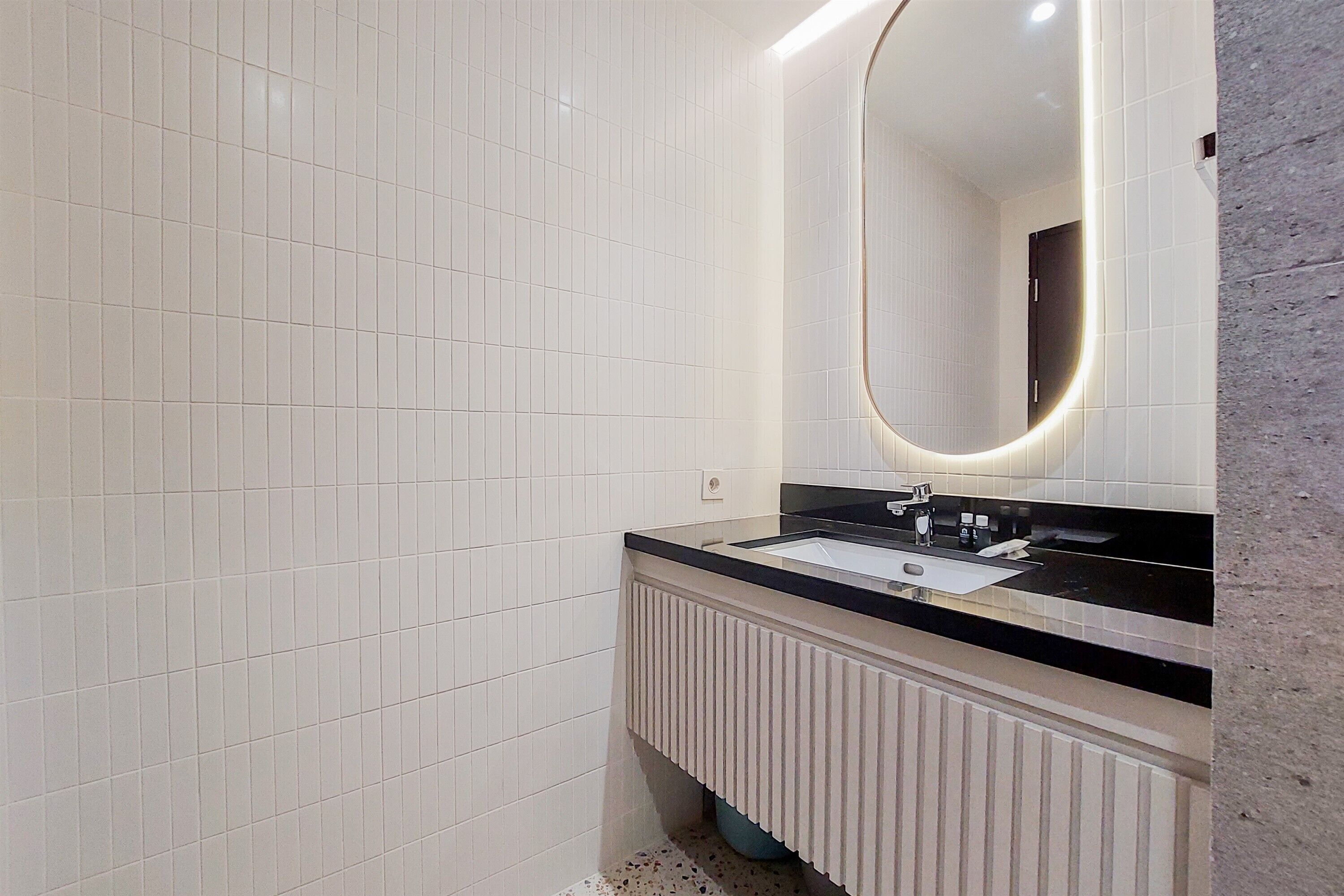 Uma casa de banho moderna com paredes revestidas a mosaico branco, bancada preta, lavatório e um espelho grande com moldura iluminada.