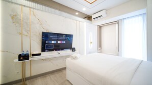 A modern hotel room with a large flat-screen TV mounted on the wall, a bed with white linens, a small bedside table with a bottle of water, and an air conditioning unit on the wall. - Modern And Cozy Studio At Benson Supermall Mansion Apartment (Surabaya)