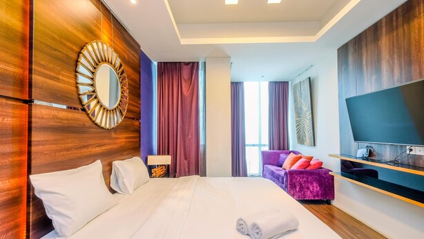 A modern hotel room with a large bed, a TV, a wooden accent wall, and a purple sofa. - Homey Penthouse 3Br With Extra Room Kemang Village Apartment (Jakarta)