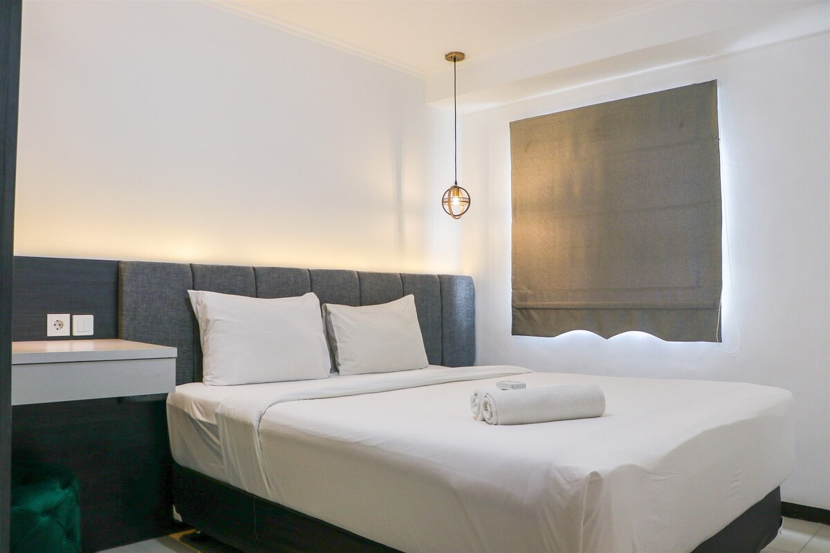 Una cama bien hecha con sábanas blancas, un cabecero gris y una mesita de noche con una lámpara.