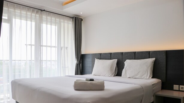 A modern hotel room with a large bed, a bedside table, and a view of the city through sheer curtains. - Spacious 3Br With Good View At Gateway Pasteur Apartment (Cimahi)