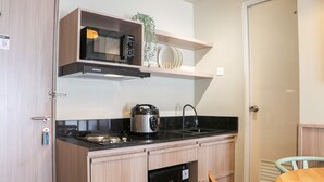 A compact kitchen with a microwave, coffee maker, and a small refrigerator. There is a wooden table and a chair, and a door with a handle. - Brand New 2Br At Tamansari Panoramic Apartment (Bandung)
