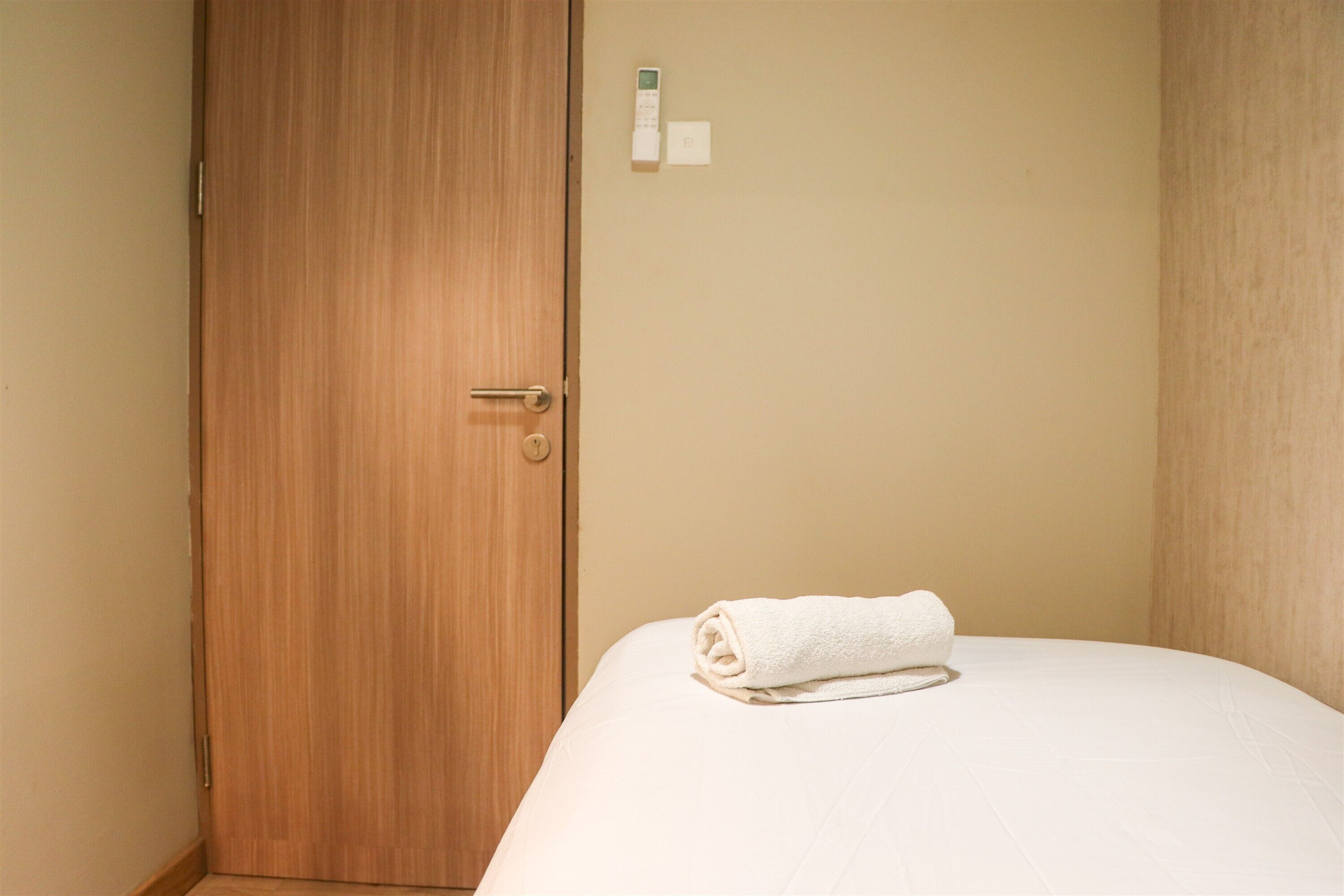 Une chambre d’hôtel avec une porte en bois, un lit blanc sur lequel est posée une serviette blanche roulée, et un thermostat fixé au mur.