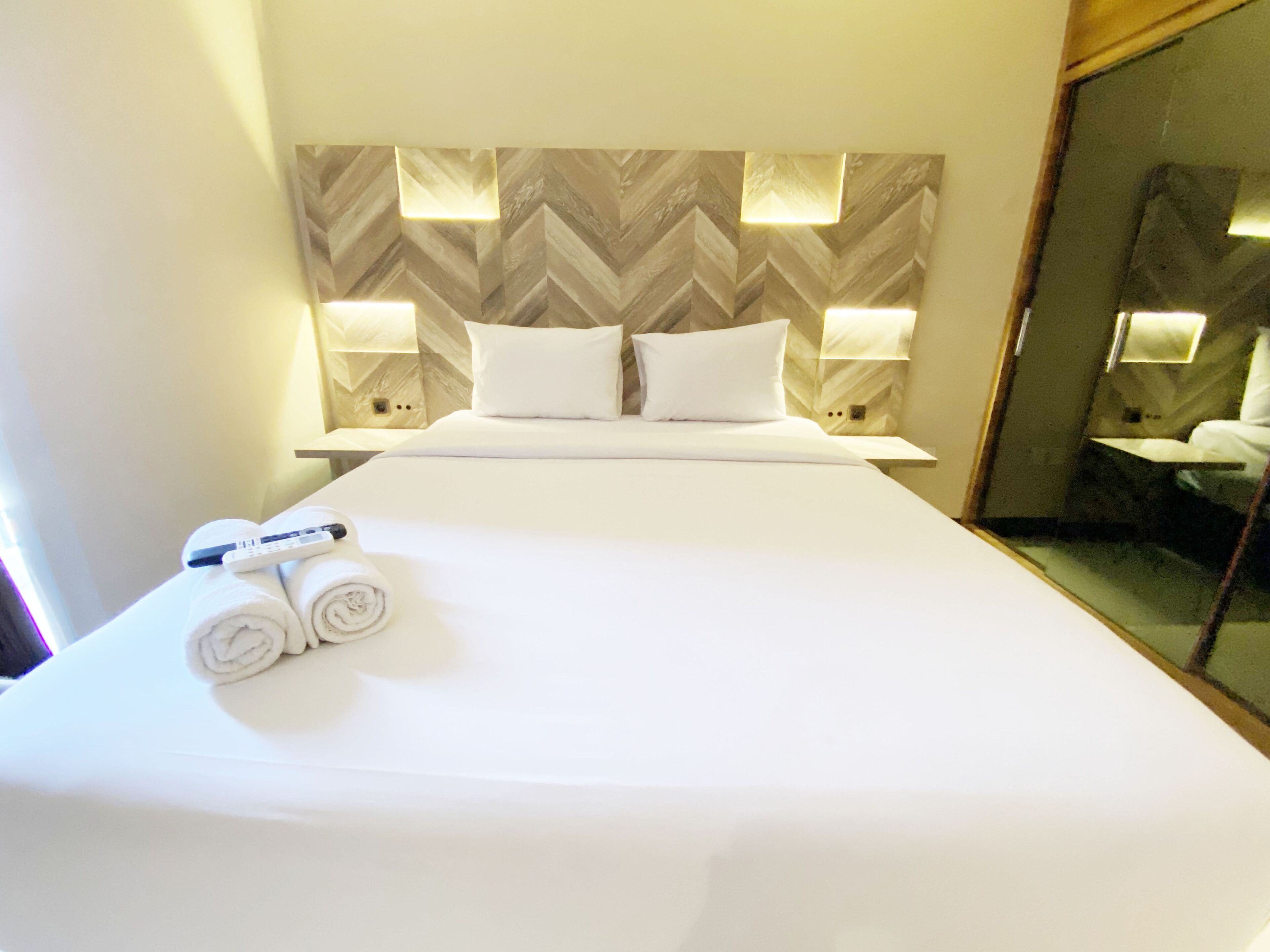 Habitación de hotel con una cama grande, ropa de cama blanca, dos almohadas, un control remoto y una toalla enrollada. Hay luces empotradas en la pared y un espejo.