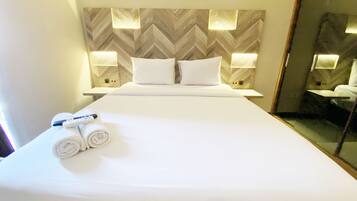 Habitación de hotel con una cama grande, ropa de cama blanca, dos almohadas, un control remoto y una toalla enrollada. Hay luces empotradas en la pared y un espejo.