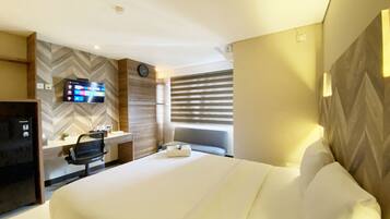 A hotel room with a bed, a desk, a chair, a TV, and a clock on the wall.