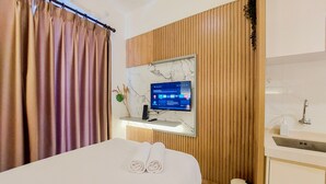 Habitación de hotel con cama, televisor montado en una pared de paneles de madera, un pequeño estante con una planta y un baño con lavabo y espejo.