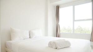 Ein großes, ordentlich bezogenes Bett mit weißen Leinen und einem zusammengerollten Handtuch an der Seite, platziert in einem hellen Raum mit einem großen Fenster und Vorhängen.