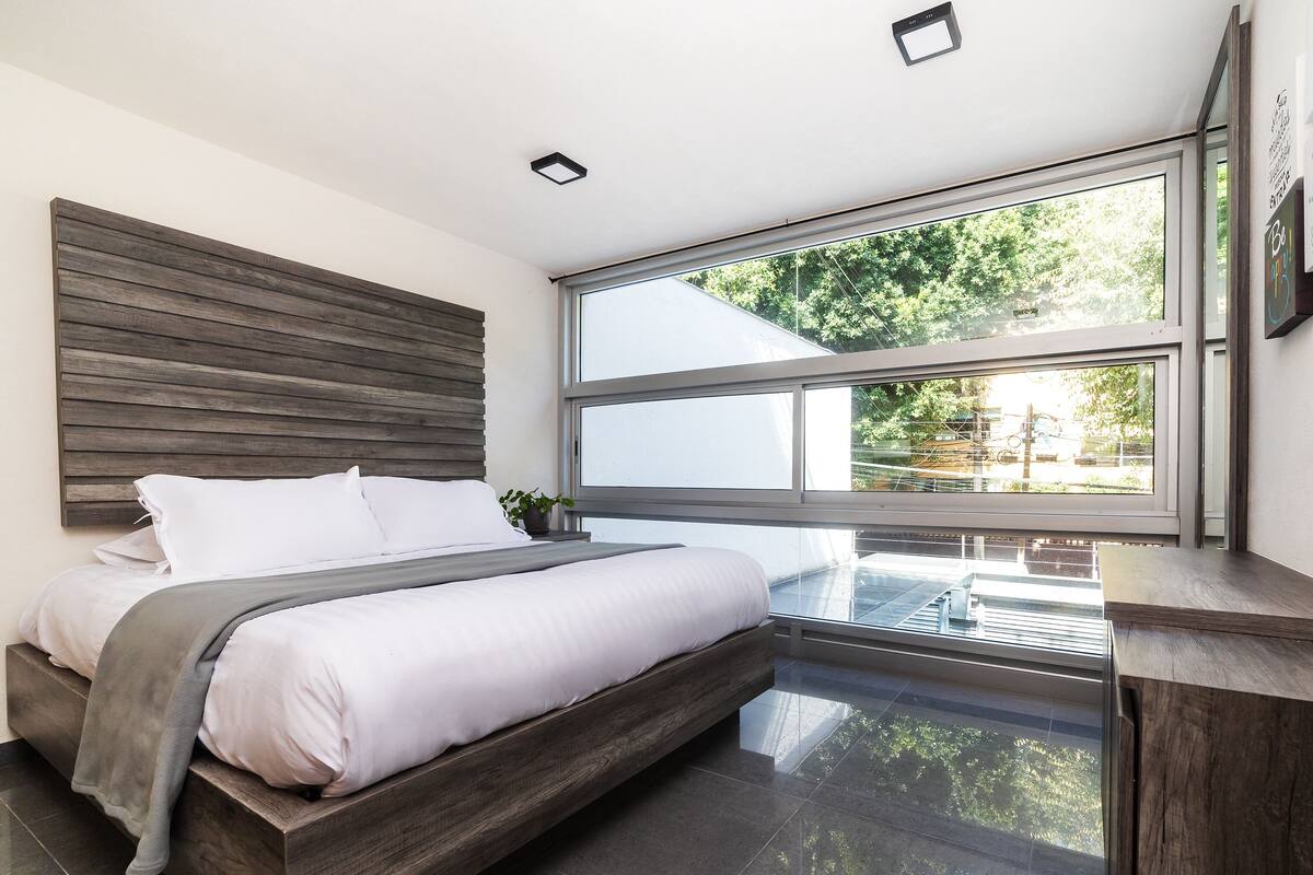 A modern bedroom with a large bed, a wooden headboard, a view of greenery outside, and a large window.