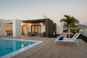 A modern outdoor pool area with a stone wall, lounge chairs, and a seating area with sofas and a table. - Villa Iris  (Playa Blanca)