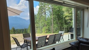 Een kamer met een groot raam met uitzicht op een grasveld met bomen en bergen. Er staat een houten tafel met stoelen en een zwarte leren bank.