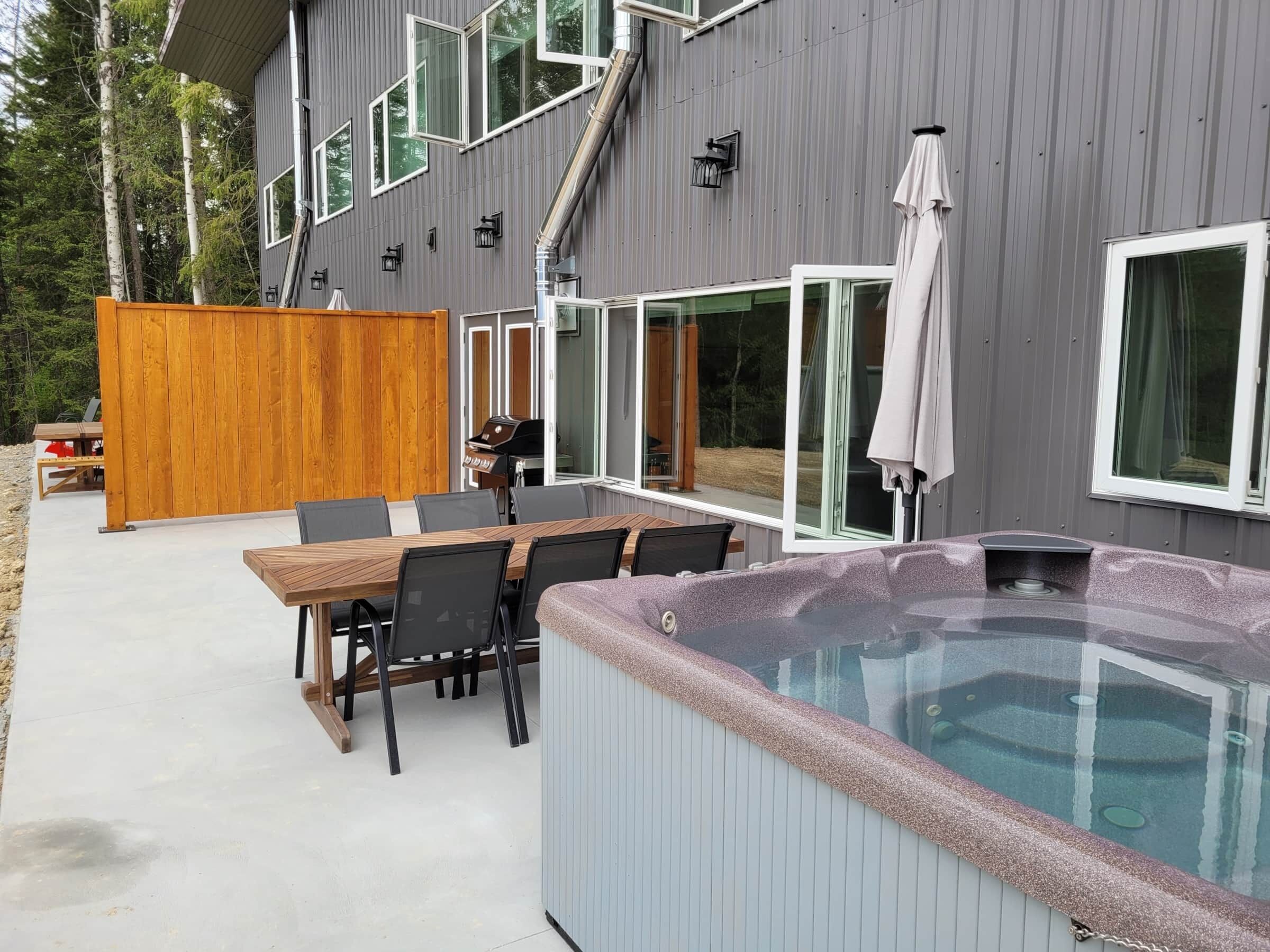 An outdoor patio with a hot tub, a wooden table, and chairs.