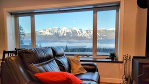 Een woonkamer met een leren bank, een rood kussen en een uitzicht op besneeuwde bergen door een groot raam.
