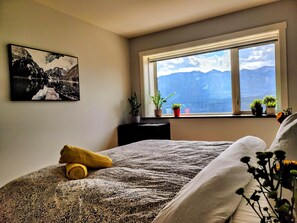 A bedroom with a bed, a window with a mountain view, a painting on the wall, and a potted plant on the windowsill.