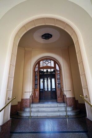 Un grand escalier menant à une porte en bois dotée de panneaux de verre, flanqué de marches en marbre et surmonté d’un luminaire circulaire au plafond.