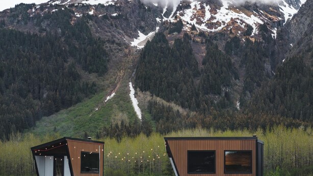 Two modern cabins with large windows, one with a slanted roof, set against a mountainous backdrop.