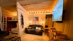 A cozy cabin interior with a wooden floor, a leather sofa, a TV mounted on the wall, and a fireplace with wood logs.