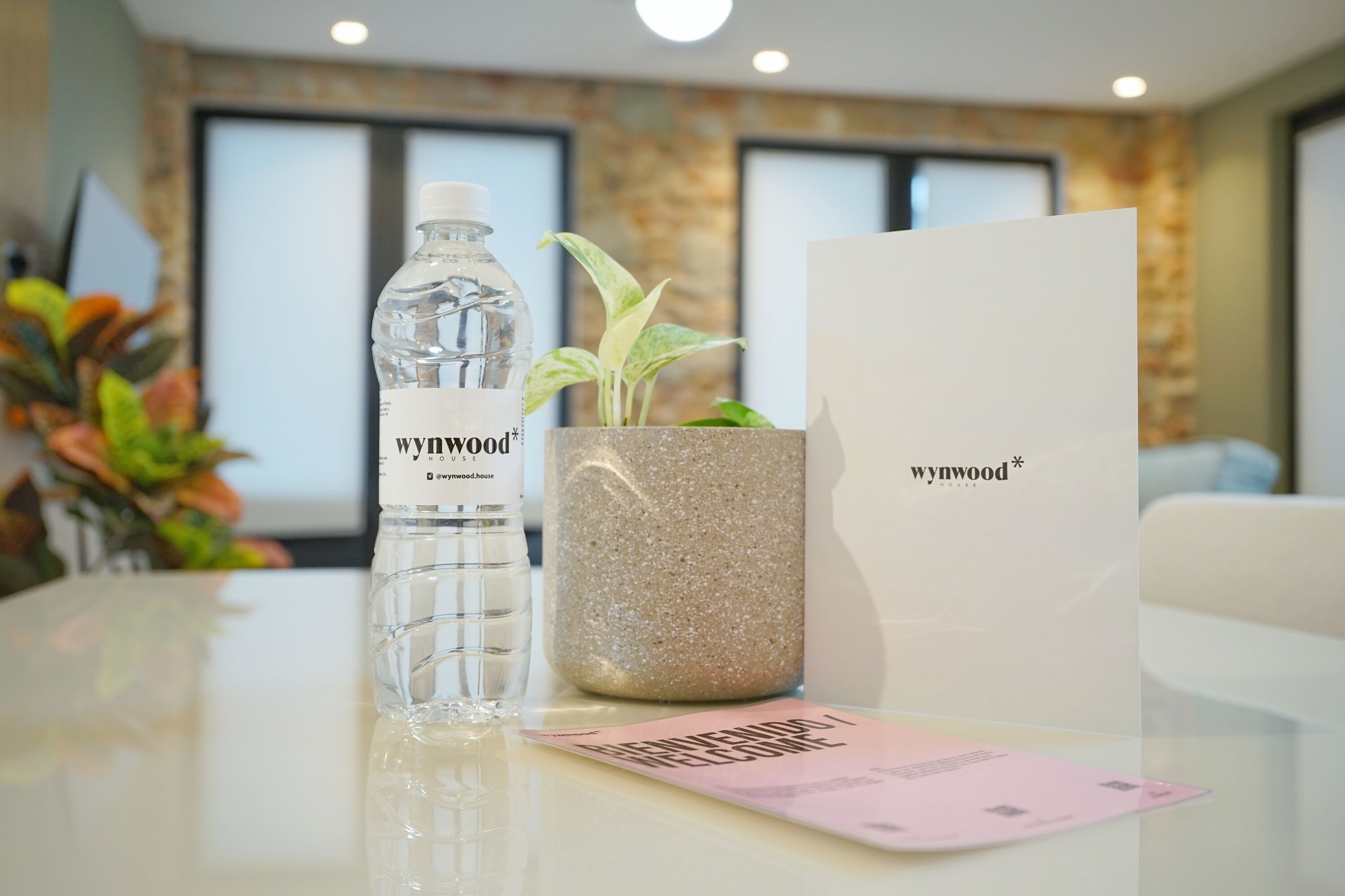 Eine klare Wasserflasche, ein Blumentopf und eine Willkommenskarte stehen auf einem Tisch in einem modernen Zimmer mit einer Steinwand und großen Fenstern.