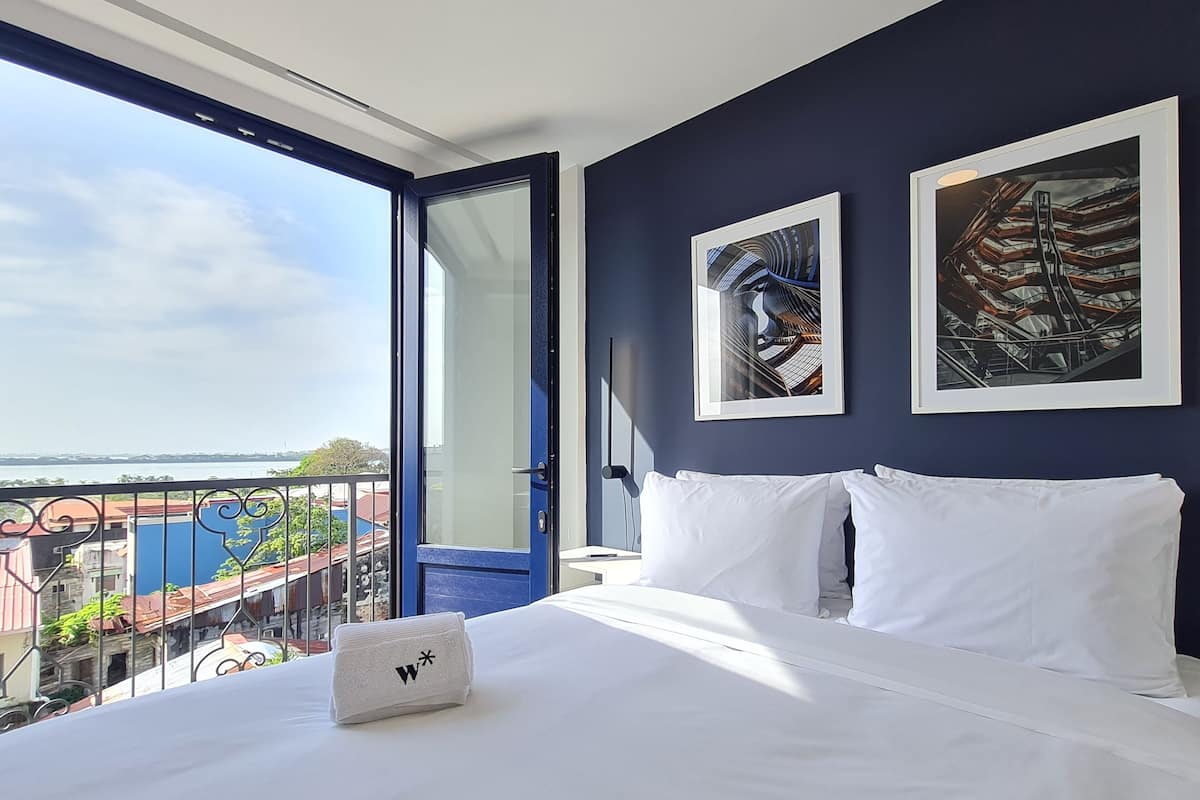 A hotel room with a large bed, two white pillows, a white towel, and two framed artworks on the wall.