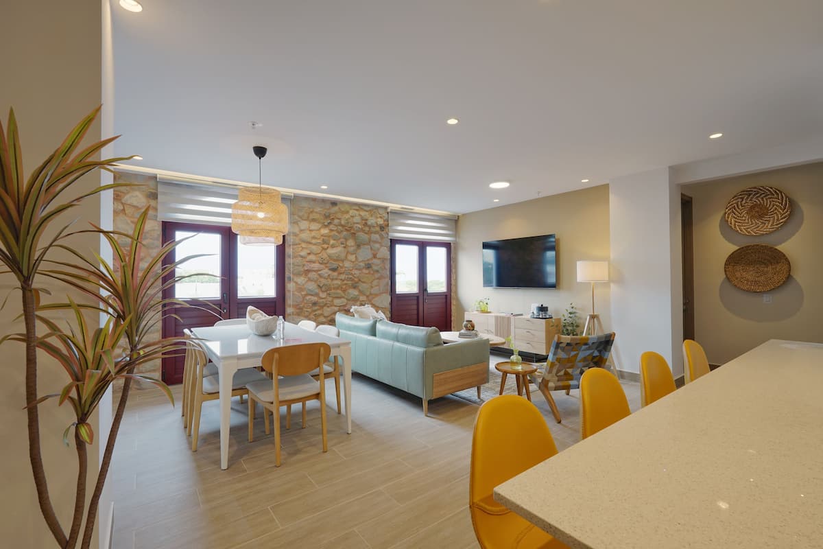 A modern living room with a sofa, dining table, and chairs. There is a stone wall and a television mounted on the wall.