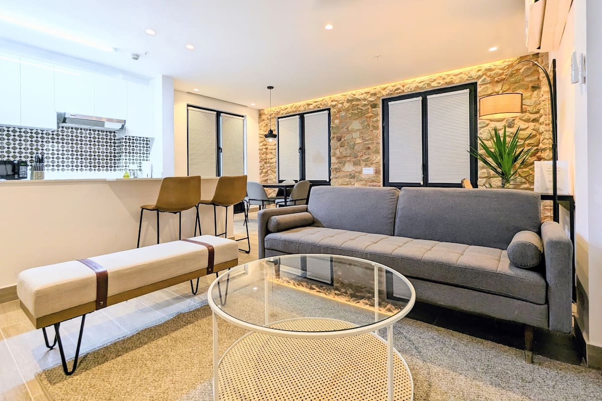 A modern living room with a grey sofa, a glass coffee table, and a bench. There is a kitchen area in the background with a stone wall and a dining table with chairs.