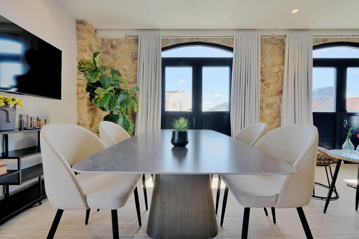 A modern dining area with a large table, six chairs, a potted plant, and a TV on the wall.