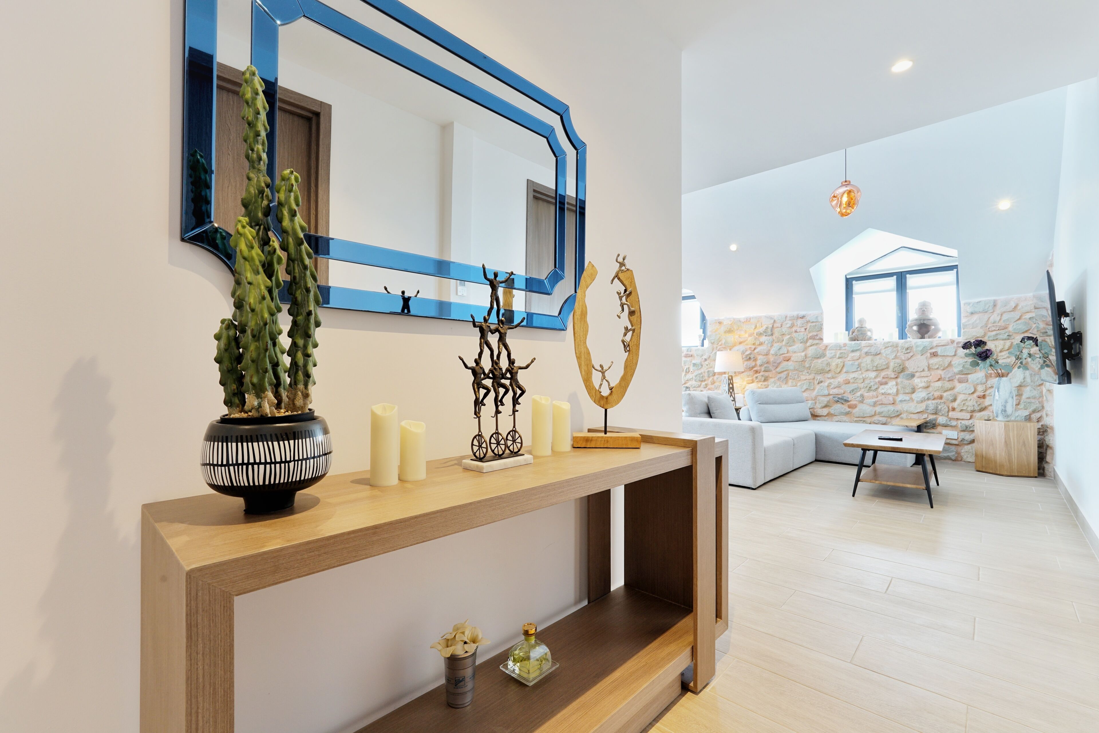 Uma sala de estar moderna com uma mesa consola de madeira, uma planta em vaso, velas e objetos decorativos. Um espelho grande com moldura azul está fixado na parede.