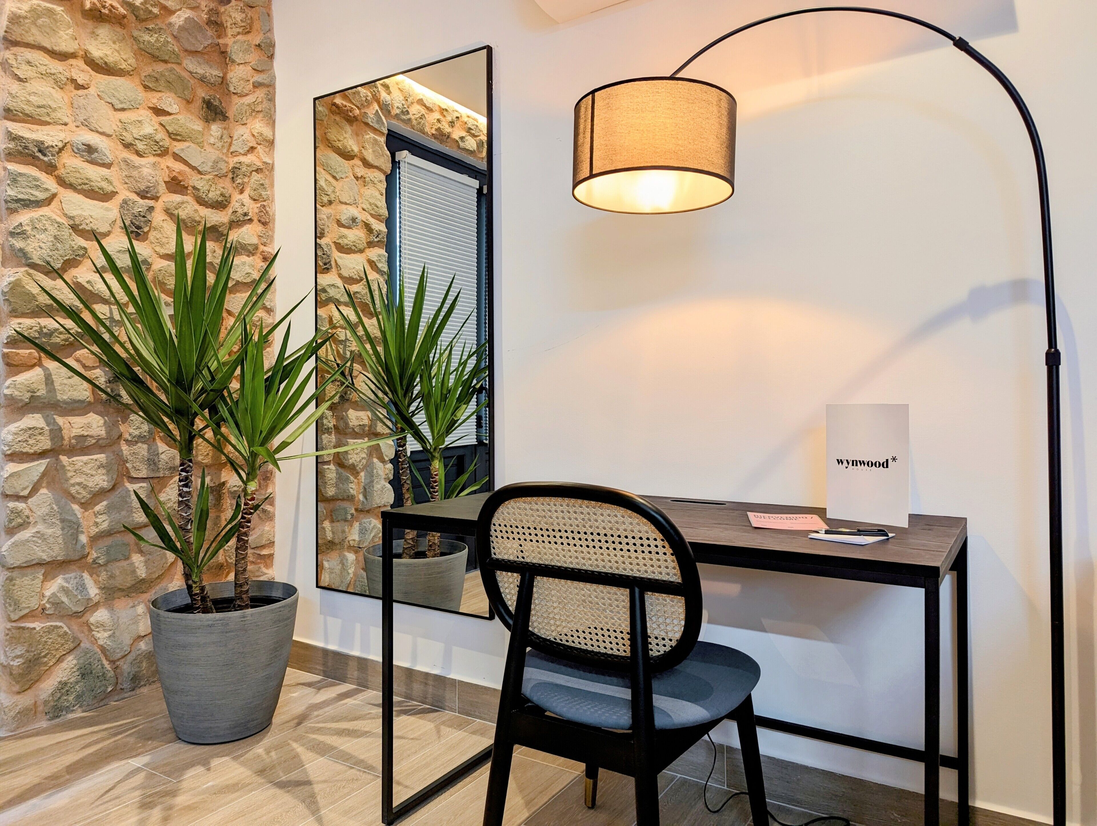 A modern room with a stone wall, a large mirror, a desk with a chair, and a floor lamp.