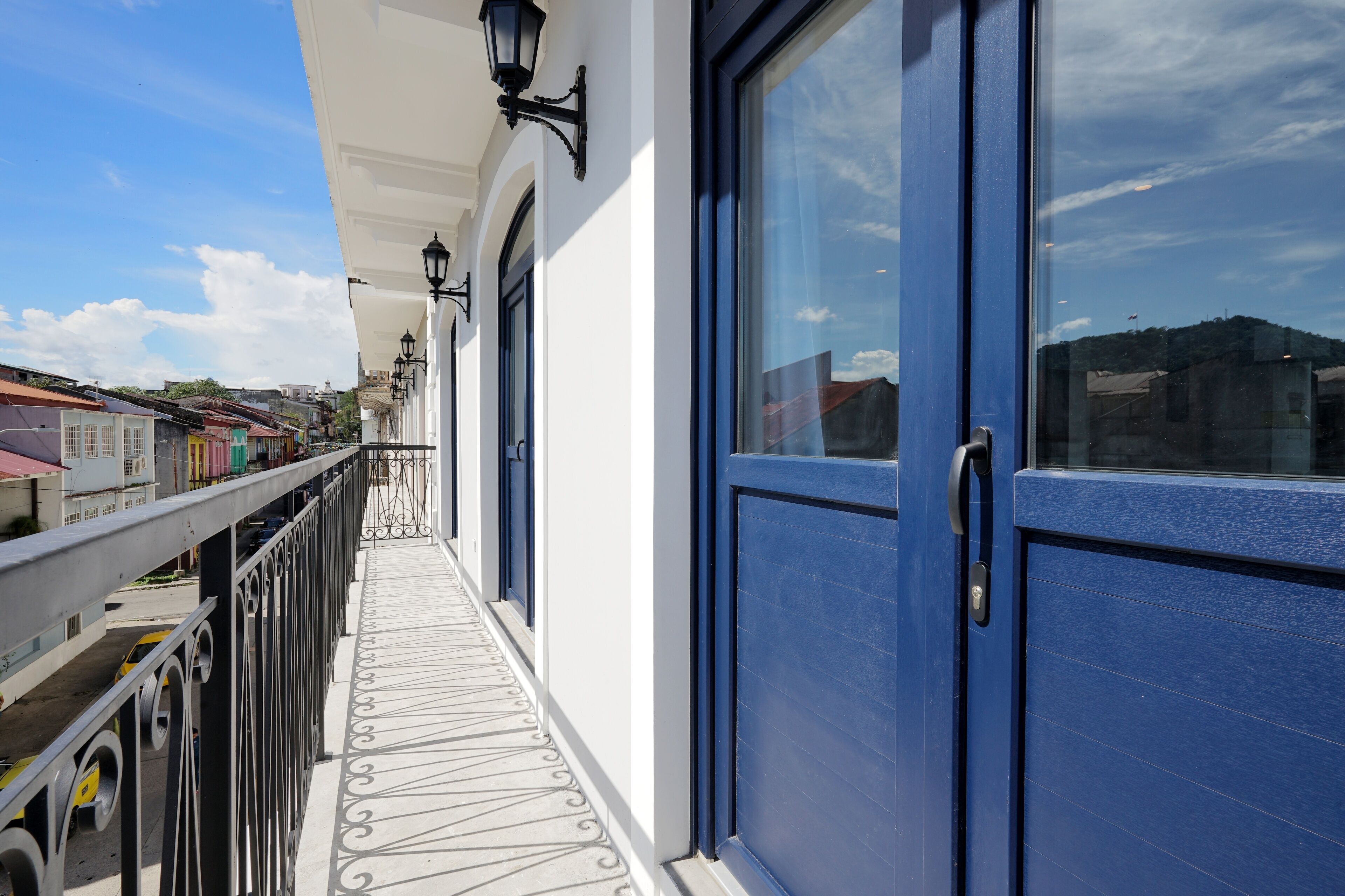 Un balcon avec une balustrade en métal, une porte bleue et un mur blanc orné d’une lanterne noire.