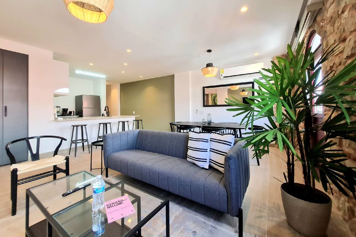 A modern living room with a grey sofa, a glass coffee table with a water bottle, and a 'Welcome' sign. There is a kitchen in the background with a bar area.