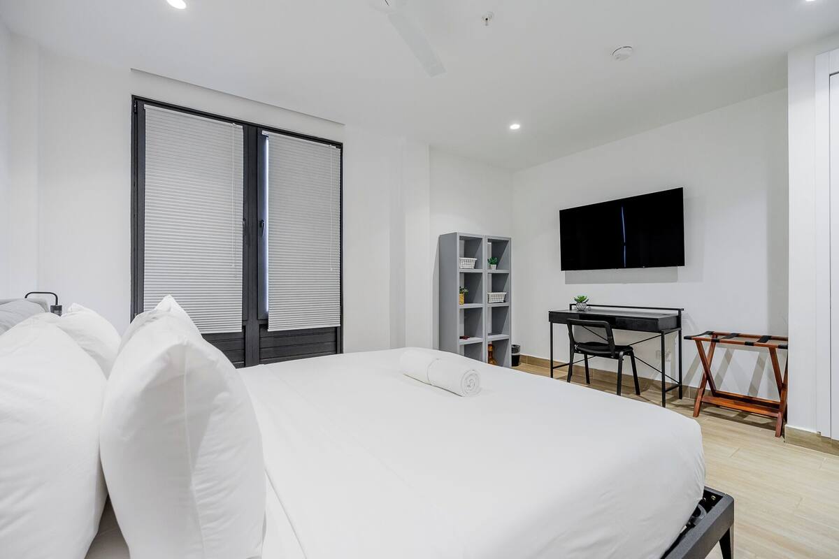 A modern hotel room with a bed, a desk, a chair, a TV, and a bookshelf.