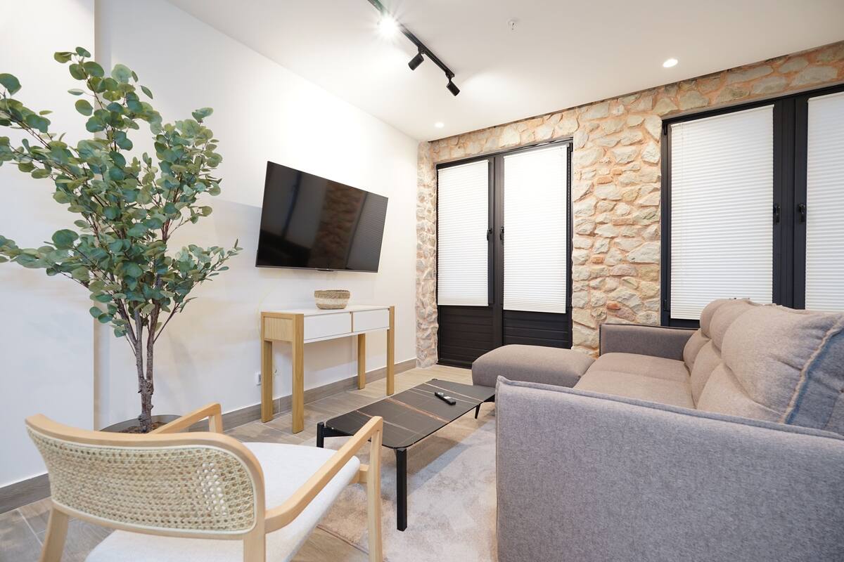 A living room with a sofa, a coffee table, a TV mounted on the wall, a stone wall, and a potted plant.