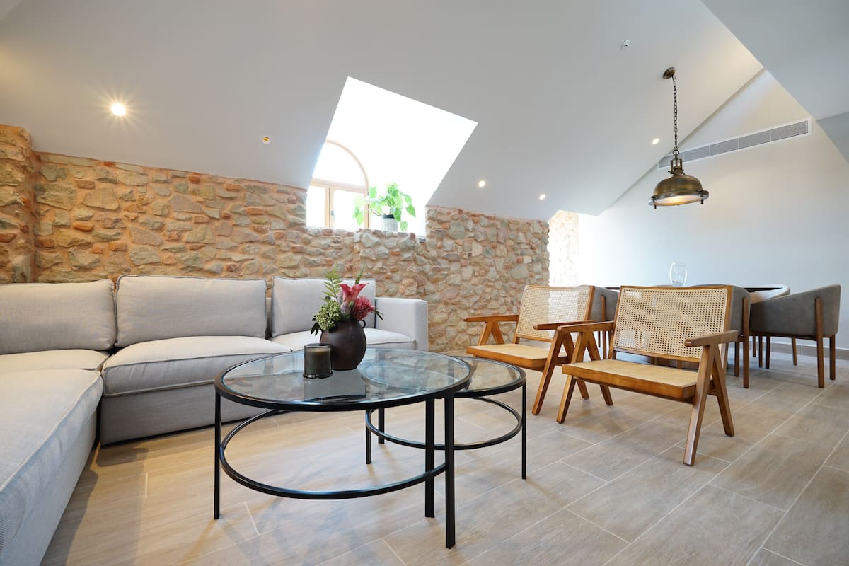 A modern living room with a stone wall, a glass coffee table, a sofa, and a dining area with chairs and a table.