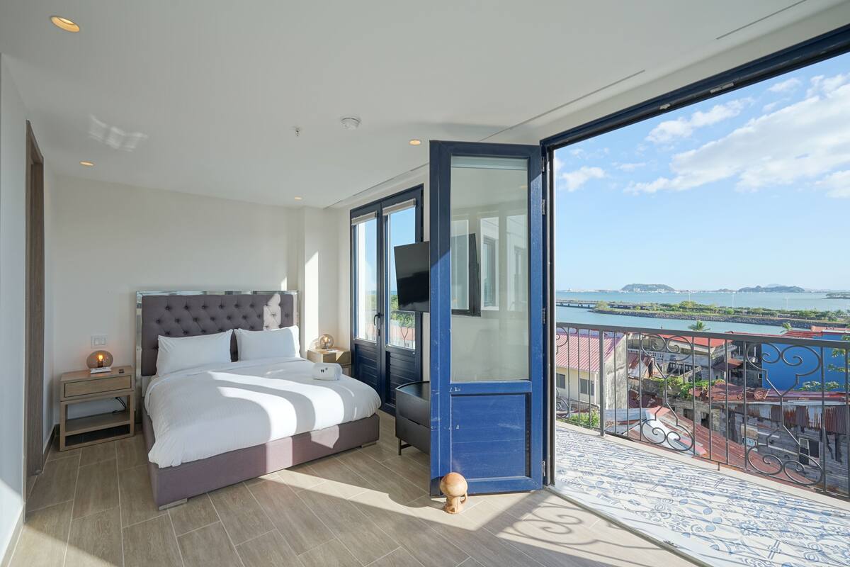 A modern bedroom with a large bed, a nightstand, a TV, and a balcony with a view of the city and water.
