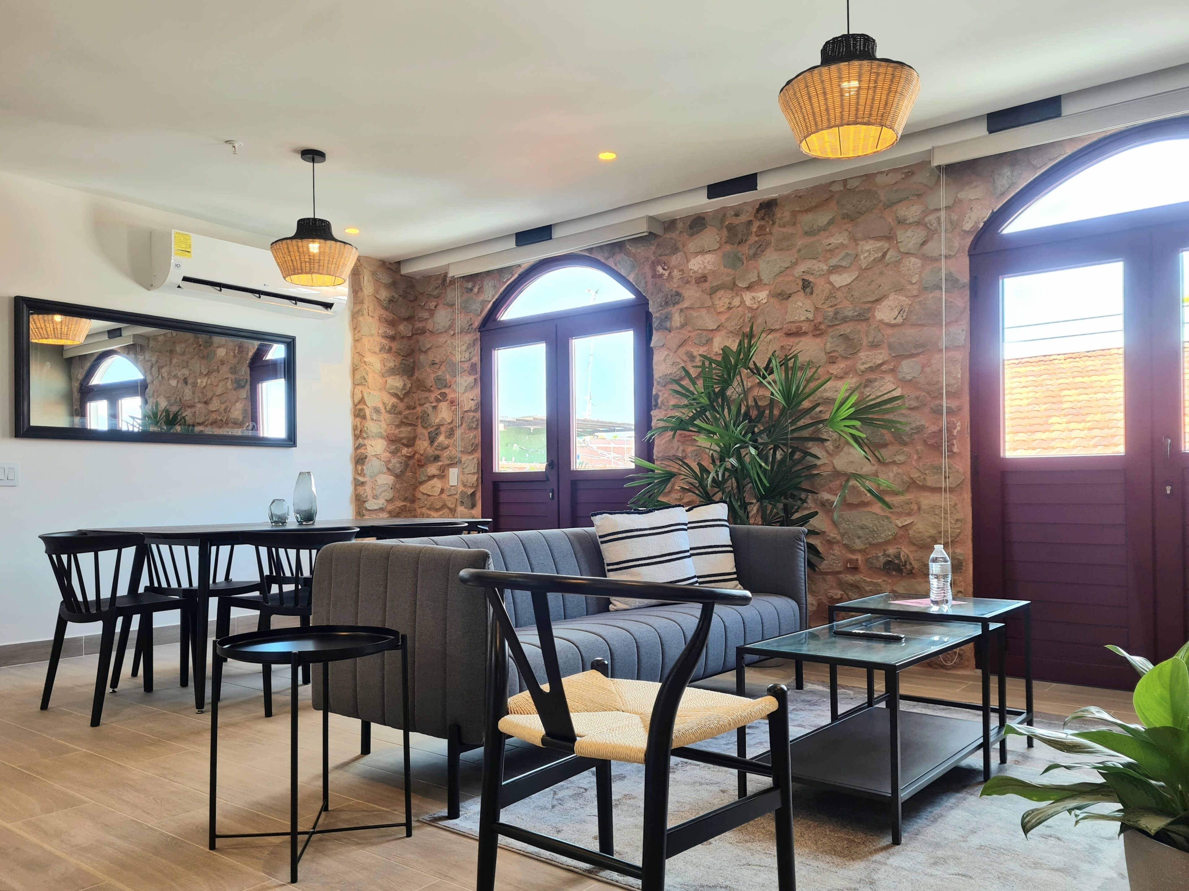 Un moderno salón con una pared de piedra, un sofá, una mesa de comedor y sillas. Hay dos ventanas grandes con arcos, una lámpara colgante y una planta en maceta.