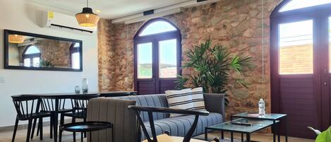 Un moderno salón con una pared de piedra, un sofá, una mesa de comedor y sillas. Hay dos ventanas grandes con arcos, una lámpara colgante y una planta en maceta.